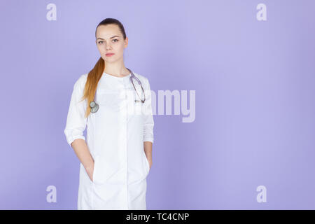 Young female blonde doctor with her hands in pockets with stethoscope on her neck on purple background with space for text. Doctod looks at camera Stock Photo