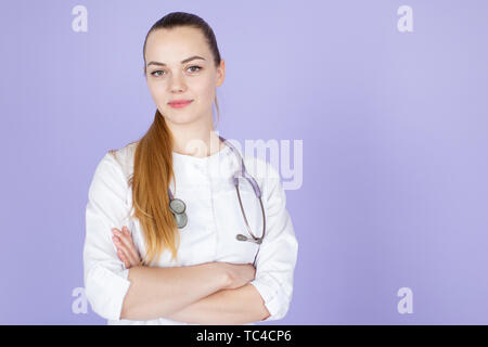 Young female blonde doctor with crossed hands with stethoscope on her neck on purple background with space for text. Doctod looks at camera Stock Photo