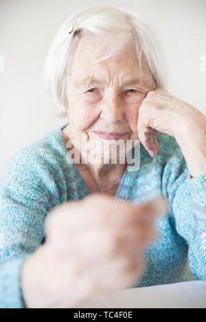 Sad elderly woman sitting at the table at home and looking miserably at only remaining coin from pension in her hand. Stock Photo