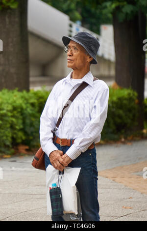 Older, mature, Japanese man wearing glasses, hat, denim jeans, leather belt, and white striped button shirt, and thinking while holding shopping bag. Stock Photo
