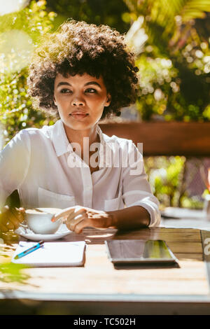 Female doing her work from outdoor coffee shop. African female sitting at a coffeeshop table and looking away thinking. Stock Photo