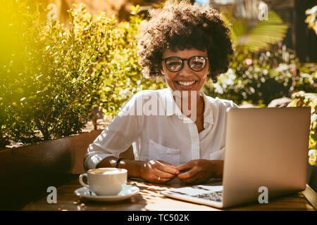 Young african woman sitting at cafe table with laptop and cup of coffee. Female with eyeglasses at coffee shop looking at camera and smiling.