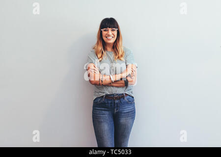 Smiling businesswoman standing against the wall with arms crossed. Cheerful woman in jeans trousers and tshirt standing with folded hands. Stock Photo