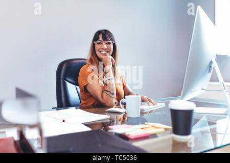 Smiling businesswoman sitting at her desk in office with hand to her chin. Cheerful woman working on computer sitting at her desk. Stock Photo