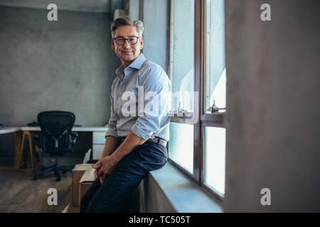 Happy mature businessman leaning by a window sill and staring to camera. Male entrepreneur at his office looking at camera. Stock Photo