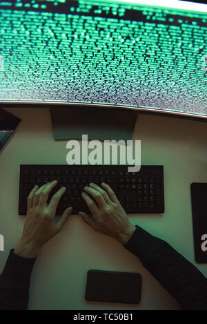 Data security. Top view of young hacker's hands stealing data while sitting in front of monitor with binary code in dark room. Cyber attack. Cyber security Stock Photo