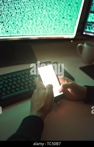 Professional Hacker. Close-up photo of young hacker in black hoodie using his smartphone for stealing data while sitting in dark room. Binary code. Cyber attack. Cyber security Stock Photo