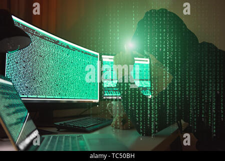 Cyber crime. Side view of young hacker in black hoodie using multiple computers and his smartphone for stealing data while sitting in dark room. Binary code. Cyber attack. Cyber security Stock Photo