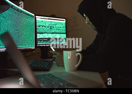 Computer security. Young man in black hoodie using multiple computers and his smartphone for stealing data while sitting in dark room. Binary code. Cyber attack. Cyber security Stock Photo