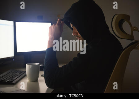Computer hacker. Young man in black hoodie using multiple computers for stealing data while sitting in dark room. Binary code. Cyber attack. Cyber security Stock Photo