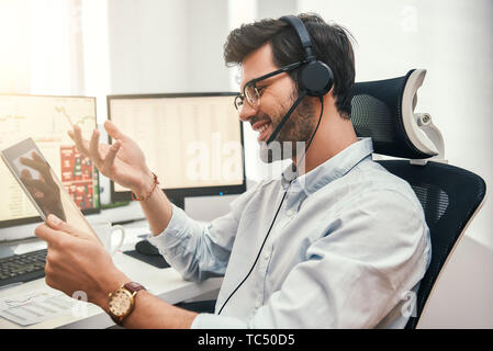 Happy trader. Successful bearded businessman in headset holding digital tablet, talking with client and smiling while sitting in front of monitor screens in the office. Digital technologies. Trade concept. Call center Stock Photo