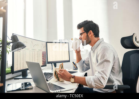 Coffee break. Young bearded businessman or trader is drinking a coffee and eating a sandwich while looking at monitor screen with financial data in his modern office. Food concept. Business concept. Trade concept Stock Photo