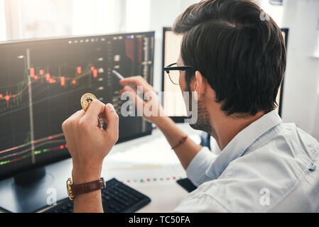 Exchange stock market. Back view of young professional trader in eyeglasses pointing on the data on computer screen with pen and holding Bitcoin in one hand while working his modern office. Crypto-currency. Virtual currency. Investment concept Stock Photo