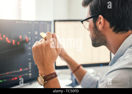 Investing. Close-up of young bearded trader in eyeglasses working with graph and financial reports on computer screen and holding Bitcoin in one hand while working his modern office. Crypto-currency. Virtual currency. Investment concept Stock Photo