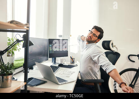 Feeling exhausted. Tired young beard businessman or trader in formal wear keeping eyes closed and stretching while sitting at his working place. Stock broker. Forex market. Trade concept Stock Photo