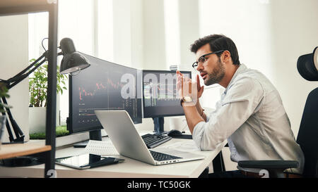 Trading strategy. Successful young trader in eyeglasses looking at analyzing trading charts on computer screens in while sitting in his modern office. Stock broker. Forex market. Trade concept Stock Photo