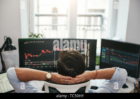 Time to relax. Back view of young businessman or trader holding hands behind head while looking at charts and financial data on computers in his modern office. Forex market. Trade concept. Investment concept Stock Photo