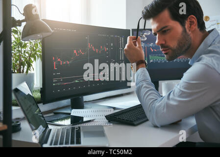 Successful investor. Serious young male trader holding eyeglasses and analyzing data and graphs on his laptop while working in his modern office. Stock exchange. Trade concept. Investment concept Stock Photo
