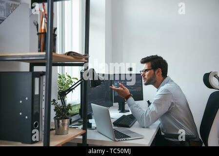 Really Side view of surprised young male stock market broker analyzing a graphs on computer and gesturing while sitting in his modern office. Stock exchange. Trade concept. Investment concept Stock Photo