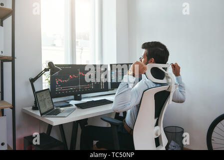 I need to relax. Sleepy young businessman or trader with hands raised feeling tired after working all day with data and charts on computers at his modern office. Forex market. Trade concept. Investment concept Stock Photo