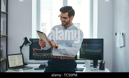 Good news. Happy young trader in formal wear is looking is using his digital tablet and smiling while standing in front of computer screens with trading charts in the office. Forex market. Trade concept. Investment concept Stock Photo
