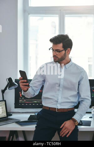 Successful businessman. Concentrated young trader in formal wear and eyeglasses is looking at trading graphs and reports on his smartphone for investment purpose while working at his office. Forex market. Trade concept. Investment concept Stock Photo