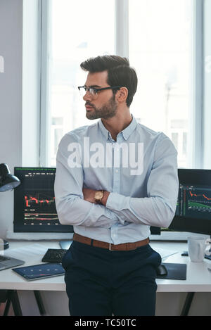 Young and smart. Vertical photo of confident businessman or trader in formal clothes and eyeglasses looking away and keeping arms crossed while standing at office. Stock broker. Forex market. Trade concept