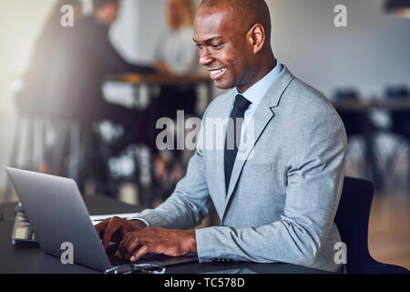 Smiling young African American businessman sitting at his desk in an office using a laptop with coworkers in the background Stock Photo