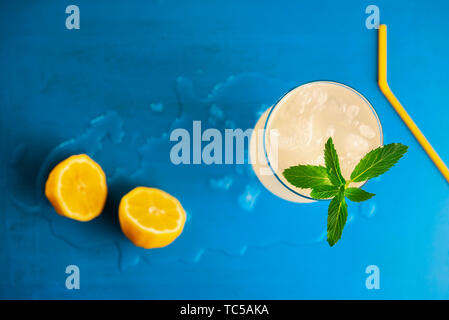 Above view of summer cold drink, a glass of lemonade with crushed ice and peppermint leaves, lemons halves and yellow straw, on a blue background. Stock Photo
