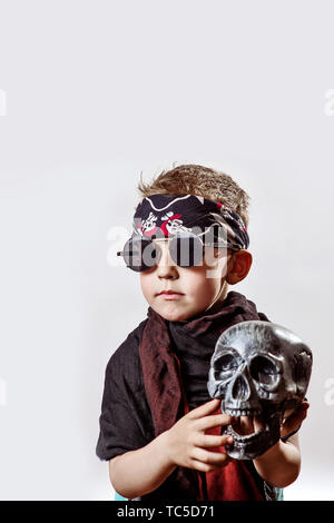 boy rocker in black glasses, scarf, bandana and with a skull in his hands on a light background Stock Photo