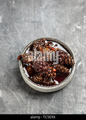 Delicious jam with baby pine cones in small bowl. Traditional Siberian dessert - young pine cones jam on gray textured background. Top view or flat lay. Stock Photo