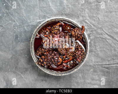 Delicious jam with baby pine cones in small bowl. Traditional Siberian dessert - young pine cones jam on gray textured background. Top view or flat lay. Stock Photo