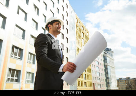 Low angle portrait of handsome Middle-Eastern businessman holding plans standing against apartment building on construction site, copy space Stock Photo