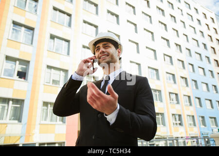Low angle portrait of handsome Middle-Eastern businessman speaking by phone standing against apartment building on construction site, copy space Stock Photo