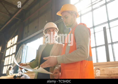 Low angle portrait of female production manager discussing plans with worker standing in indusrial workshop lit by sunlight Stock Photo