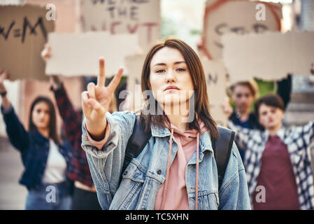 We want peace. Young woman is showing piece sign while standing in front of female activists holding signboards. Women rights. Women's march. Human rights Stock Photo