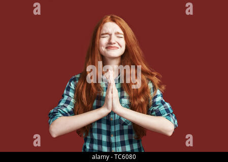 Studio shot of redhead girl in checkered shirt worrying, holding hands in pray, apologizing for bad behaviour or mistake, standing over red background with closed eyes Stock Photo