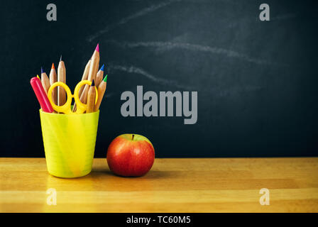 Colored pencils in yellow cup and fresh apple on the table on chalkboard background. Stock Photo