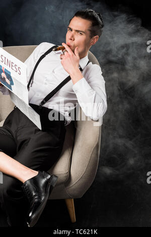 overhead view of handsome man sitting in armchair holding business newspaper and smoking cigar on black with smoke Stock Photo