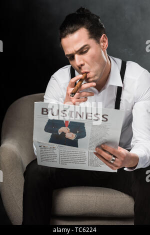 serious man sitting in armchair reading business newspaper and smoking cigar on black with smoke Stock Photo