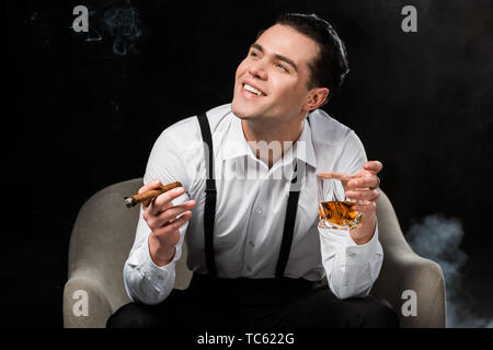 happy man sitting in armchair and holding glass of whiskey and cigar on black with smoke Stock Photo