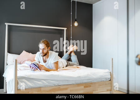 Handsome man in pajamas reading book while relaxing on the bed at home Stock Photo