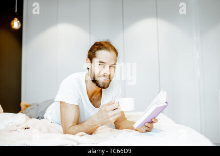 Handsome man in pajamas reading book while relaxing with coffee on the bed at home Stock Photo