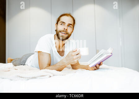 Handsome man in pajamas reading book while relaxing with coffee on the bed at home Stock Photo