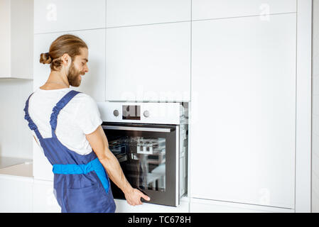 Handsome workman in workwear installing electric oven into the kitchen shelves at the modern kitchen at home Stock Photo