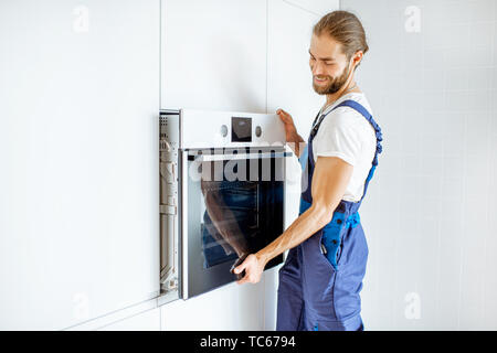 Handsome workman in workwear installing electric oven into the kitchen shelves at the modern kitchen at home Stock Photo