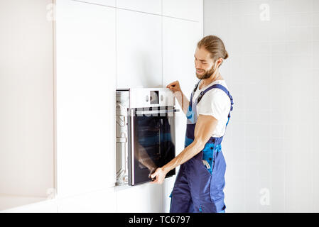 Handsome workman in workwear installing electric oven into the kitchen shelves at the modern kitchen at home Stock Photo