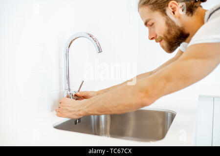 Handsome workman fixing or installing faucet in the white interior kitchen at home Stock Photo