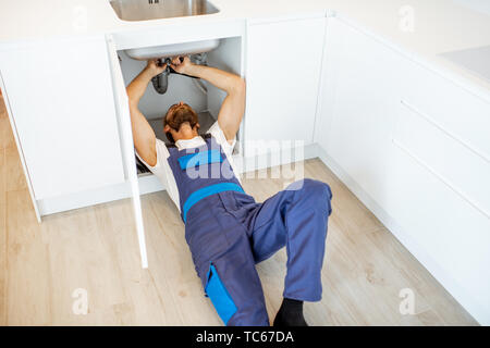 Plumber in overalls repairing or installing sewerage under the kitchen sink at home Stock Photo