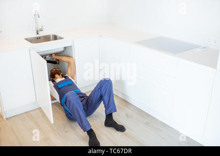 Plumber in overalls repairing or installing sewerage under the kitchen sink at home Stock Photo
